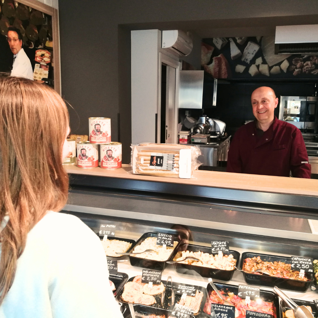 Roberto sorridente al banco della Salumeria da Adriano accoglie una cliente tra piatti pronti e specialità